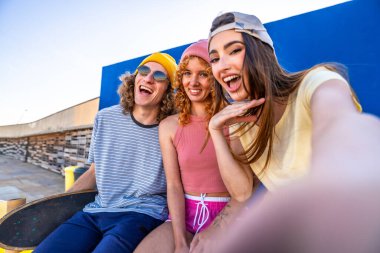 Güneşli bir günün tadını çıkaran üç neşeli patenci, canlı mavi bir duvara karşı eğlenceli bir selfie çekiyorlar, arkadaşlığı ve gençlik enerjisini kutluyorlar.
