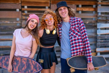 Üç genç patenci, kaykaylarıyla birlikte ahşap bir duvarın önünde gülümseyerek ve poz vererek arkadaşlıklarını ve tarzlarını sergiliyorlar.