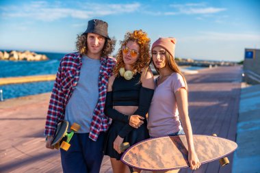 Kaykaylı üç arkadaş deniz kenarında güneşli havanın ve ahşap bir kaldırımda havanın tadını çıkarıyorlar.