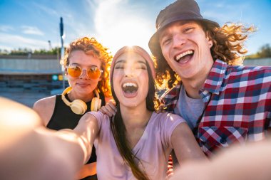 Üç neşeli genç arkadaş dışarıda selfie çekiyorlar, birlikte gülüyorlar ve güneşli bir günün tadını çıkarıyorlar.