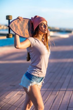 Omzunda kaykayla gezen kendine güvenen genç bir kadın. Rıhtımda güneşli bir günün tadını çıkarıyor. Aktif bir yaşam tarzı sergiliyor.