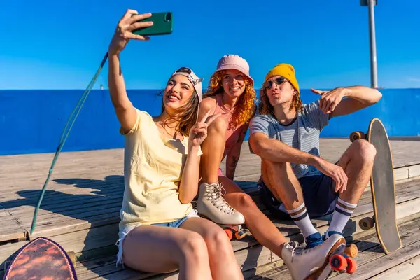 Üç genç, şık arkadaş ahşap bir tahta kaldırımda oturuyorlar, akıllı telefonlarıyla selfie çekiyorlar paten giyip güneşli havanın tadını çıkarıyorlar.