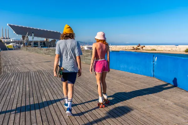 Genç bir adam kaykay gezintileri yapıyor. Tahta bir iskelede patenci bir kadının yanında. Deniz kenarında güneşli bir günün tadını çıkarıyor.