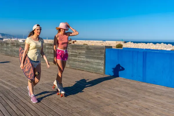 İki genç kadın deniz kenarında güneşli bir günü kucaklıyor, biri kaykay taşıyor, diğeri tahta bir kaldırımda paten kayıyor.