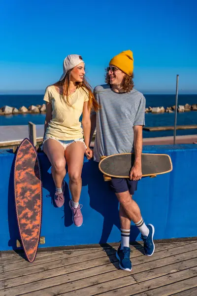 Mutlu genç patenciler deniz kenarındaki mavi bir duvarda oturuyor, ellerinde uzun kaykaylarıyla gülümsüyorlar.