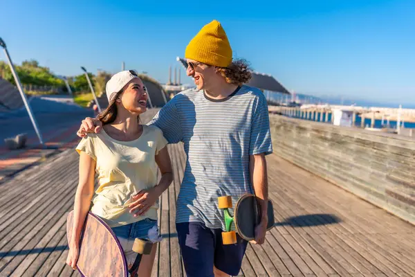 Mutlu genç çift rıhtımda el ele yürüyor, kaykay tutuyorlar ve güzel bir yaz gününde güneşin tadını çıkarıyorlar.