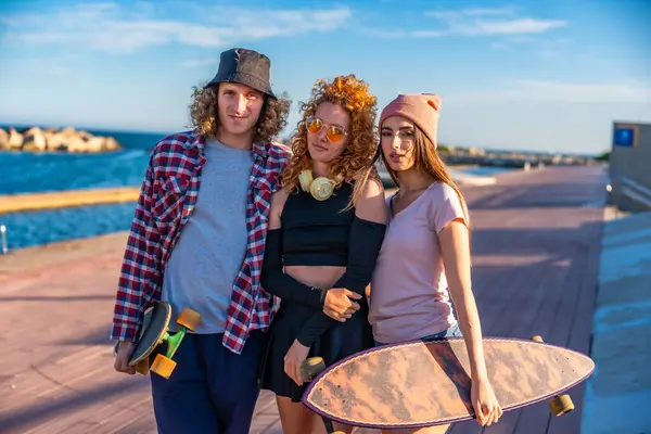 Kaykaylı üç arkadaş deniz kenarında güneşli havanın ve ahşap bir kaldırımda havanın tadını çıkarıyorlar.