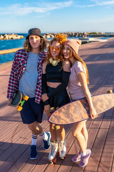 Üç mutlu kaykaycı arkadaş deniz kıyısındaki bir kaldırımda birlikte poz veriyorlar, ellerinde kaykaylar ve patenler var.