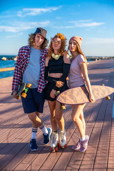 Kaykaylı üç arkadaş, güneşli bir okyanus gününün tadını çıkarıyorlar. Ahşap bir kaldırımda gündelik tarzlarını ve gençlik enerjilerini sergiliyorlar.