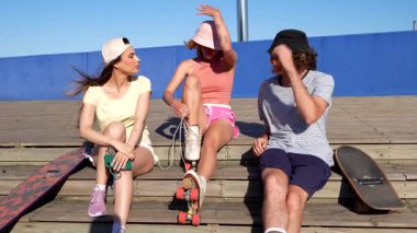 Genç arkadaşlar paten macerası için hazırlanıyorlar, heyecan paylaşıyorlar ve güneşli şehir zemininde ahşap merdivenler ve mavi duvarla ekipman hazırlıyorlar.