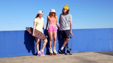 İki kız ve bir oğlan, yaz kıyafetleri içinde, kaykayları ve patenleriyle güneşli bir günde mavi bir duvara karşı sohbet ediyorlar.