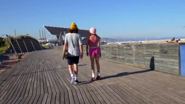 Tahta kaldırımda yürüyen mutlu genç çift, kız paten kayan ve kolunun altında kaykaylı erkek, yaz gününün tadını çıkarıyor.