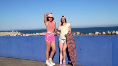 Son moda yaz kıyafetleri giymiş iki genç kadın, deniz kenarındaki canlı mavi duvara karşı kaykaylarıyla poz veriyorlar.