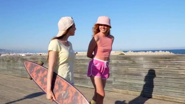 İki genç kadın güneşli bir günün tadını çıkarıyorlar, okyanus kıyısında sörf tahtaları ile yürüyorlar, sohbet ediyorlar ve gülümsüyorlar.