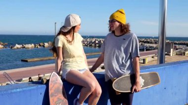 Rahat bir çift, okyanus kenarında güneşli bir günün tadını çıkarıyor, kaykaylı mavi bir duvarda oturuyor, sohbet ediyor ve sakin sahil atmosferini emiyor.