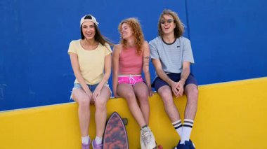 Bir grup patenci arkadaş sarı bir duvarda takılıyorlar, mavi arka planda, konuşuyorlar ve gülümsüyorlar.