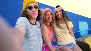 Üç neşeli arkadaş, iki kız ve bir oğlan, kaykay rampasında mavi bir duvara karşı selfie çekiyorlar.