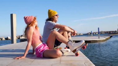 Deniz kenarındaki iskelede oturan genç patenci çift, güneşli yaz tatili boyunca neşe ve bağlantı yayıyorlar, kaygısız kıyı yaşamını kucaklıyorlar.