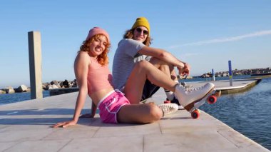 Paten giyen genç çift ahşap iskelede yan yana oturuyor güneşli bir yaz gününün keyfini çıkarıyorlar, köpüklü okyanus sularının yanında, samimi bir anı paylaşıyorlar.