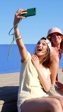 Bir grup arkadaş deniz kenarındaki ahşap iskelede otururken selfie çekiyorlar. Patenler ve günlük giysiler giyiyorlar.