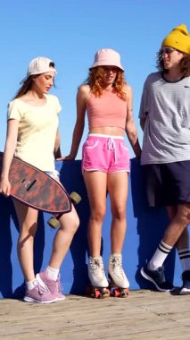 Bir grup arkadaş paten ve yaz kıyafetleri giyer, kaykay tutarlar, güneşli bir günde mavi duvarın yanında sohbet ederler.