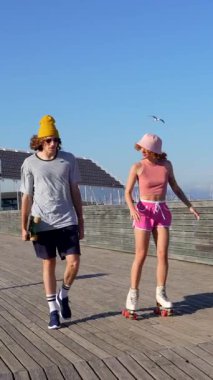 Genç çift paten kayıyor ve güneşli iskelede yürüyor, mutluluk saçıyor ve yaz tatilinin tadını çıkarıyor.