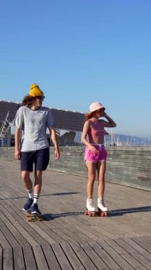 Genç çift neşeyle kaykay ve paten kayıyor. Güneşli bir tahta yolda yan yana. Açık havanın tadını çıkarıyorlar.