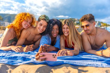 Beş neşeli genç arkadaş plaj havlusunda uzanıyorlar, akıllı telefonlarıyla selfie çekiyorlar ve yaz tatilinin tadını çıkarıyorlar.