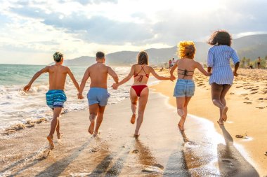 Gün batımında plajda el ele koşan, yaz tatilinin tadını çıkaran beş çok ırklı arkadaş.