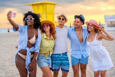 Bir grup çok ırklı arkadaş gün batımında sahilde selfie çekiyor, yaz tatilinin tadını çıkarıyorlar.