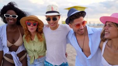 Sahilde tatilin tadını çıkaran, selfie çeken ve kumlu sahil şeridi boyunca neşeli etkileşimler paylaşan bir grup genç arkadaş.