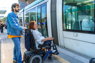 Genç adam, modern bir tramvaya binip ulaşılabilirliği ve dahil edilmeyi ön plana çıkaran tekerlekli sandalyedeki orta yaşlı kadına yardım ediyor.