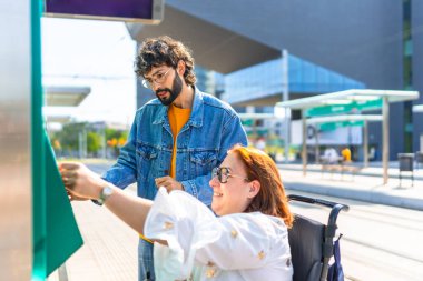 Tekerlekli sandalyedeki bir kadın modern bir tramvay durağında biletini onaylıyor. Bir arkadaşı da bu süreçte yardım ve destek sağlıyor.