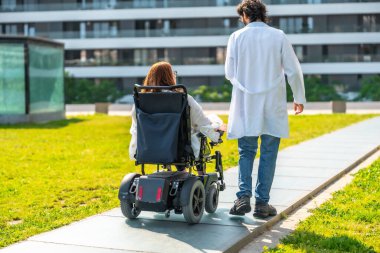 Doktor, hastaya tekerlekli sandalyede yardım ediyor güneşli bir hastane bahçesinde geziniyor iyileşme sürecini hızlandırıyor.