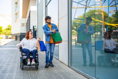 Alışveriş sonrası kaldırımda yürüyen çift tekerlekli sandalye kullanan ve renkli çantalar taşıyan kadın.