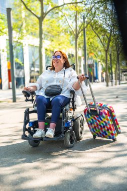 Gülümseyen kızıl saçlı turist yaz tatillerinin tadını çıkarıyor, elektrikli sandalyesine biniyor ve şehir parkında renkli valizler taşıyor.
