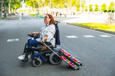 Engelli mutlu kadın, şehir caddesinde modern elektrikli tekerlekli sandalye sürerken renkli bavulunu çekerek ulaşılabilir yolculuğunun keyfini çıkarıyor.