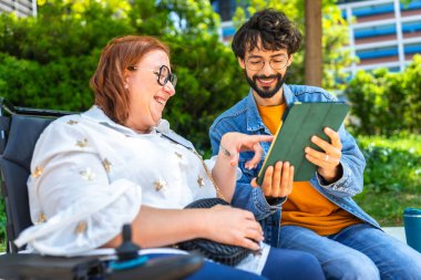 Bir adam ve bir kadın parkta dijital tablet kullanıyorlar, kadın tekerlekli sandalyede oturuyor ve ikisi de gülümsüyor.