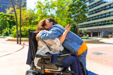 Elektrikli sandalye kullanan engelli bir kadın güneşli bir şehir parkında arkadaşından sıcak bir kucaklama alıyor.