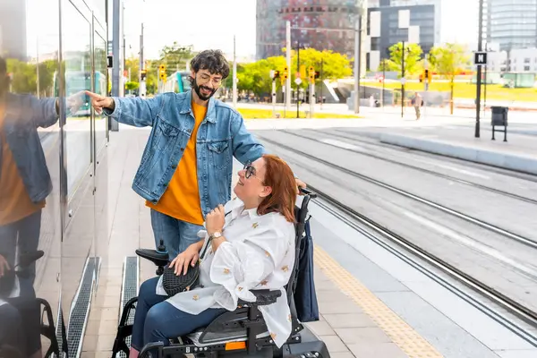Adam, modern tramvay istasyonunda elektrikli sandalye kullanan orta yaşlı bir kadınla konuşurken zaman çizelgesini işaret ediyor.