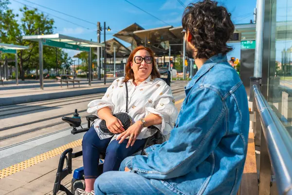 Motorlu tekerlekli sandalye kullanan bir kadın modern bir tramvay durağındaki bir adamla dostça sohbet etmekten zevk alıyor.