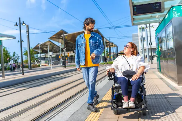 Adam, modern bir tramvay ya da tren istasyonunda beklerken elektrikli sandalye kullanan bir kadınla yürüyor ve konuşuyor.