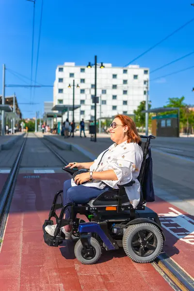 Elektrikli sandalye kullanan gülümseyen kadın güneşli bir günde modern bir tramvay istasyonunda bekliyor.