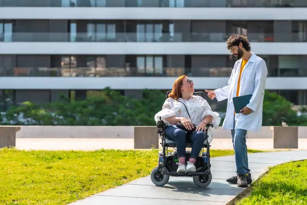 Doktor tekerlekli sandalyedeki gülümseyen engelli kadınla konuşuyor. Hastanenin yakınındaki şehir parkında güneşli bir günün tadını çıkarıyor. Erişilebilirliği ve sağlık hizmetlerini destekliyor.