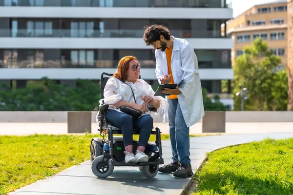 Elinde tablet olan doktor, şehir parkında tekerlekli sandalyede oturan gülümseyen kadına teşhisi açıklıyor.