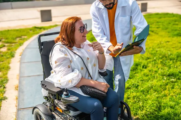 Doktor, parkta tekerlekli sandalyede oturan gülümseyen bir kadına tıbbi bilgi veriyor. Erişilebilirliği ve sağlık desteğini destekliyor.