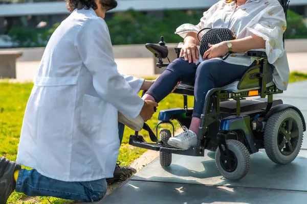 Doktor, elektrikli sandalyede oturan bir hastanın ayak bilekliğini düzeltiyor. Park ortamında kişisel bakım ve destek sağlıyor.