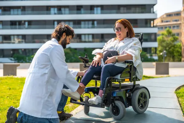 Erkek doktor, gülümseyen bir kadın için elektrikli sandalyenin en üst basamağını ayarlıyor. Güneşli bir park ortamında ulaşılabilirlik ve sağlık hizmetlerini teşvik ediyor.