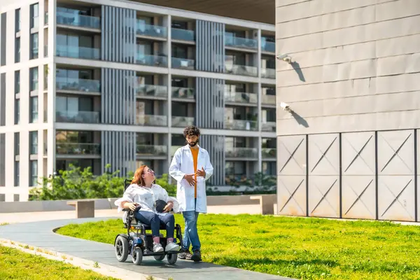 Doktor tekerlekli sandalyedeki bir hastaya yardım ediyor güneşli bir hastane bahçesinde geziniyor, hareket kabiliyetini ve refahını arttırıyor.