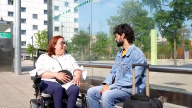 Tekerlekli sandalye kullanıcısı ve yoldaşı otobüs durağındaki canlandırıcı sohbeti paylaşıyor, yaz mevsiminde kapsayıcı dostluk ve pozitif sosyal bağlantı sergiliyor